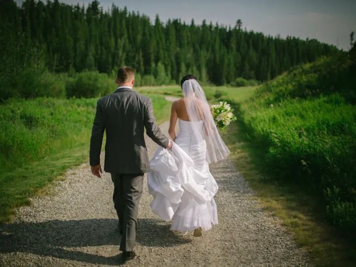 Bride and groom walking away on a gravel path toward evergreen forest, groom holding her train