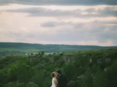 Bride and groom kissing in the lower frame against rolling green hills and cloudy sky
