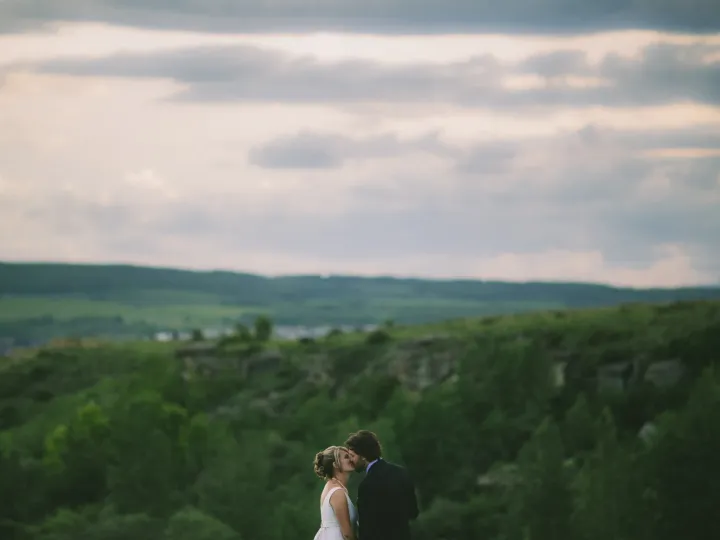 Bride and groom kissing in the lower frame against rolling green hills and cloudy sky