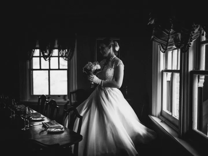 Black and white photo of a bride in gloves and a full gown standing between bright windows