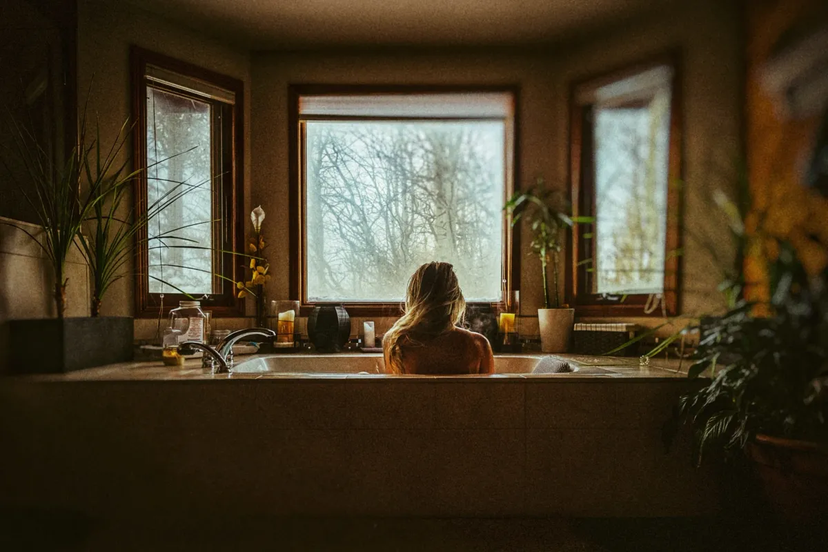 Person sitting in a bathtub beneath warm window light in a rustic room