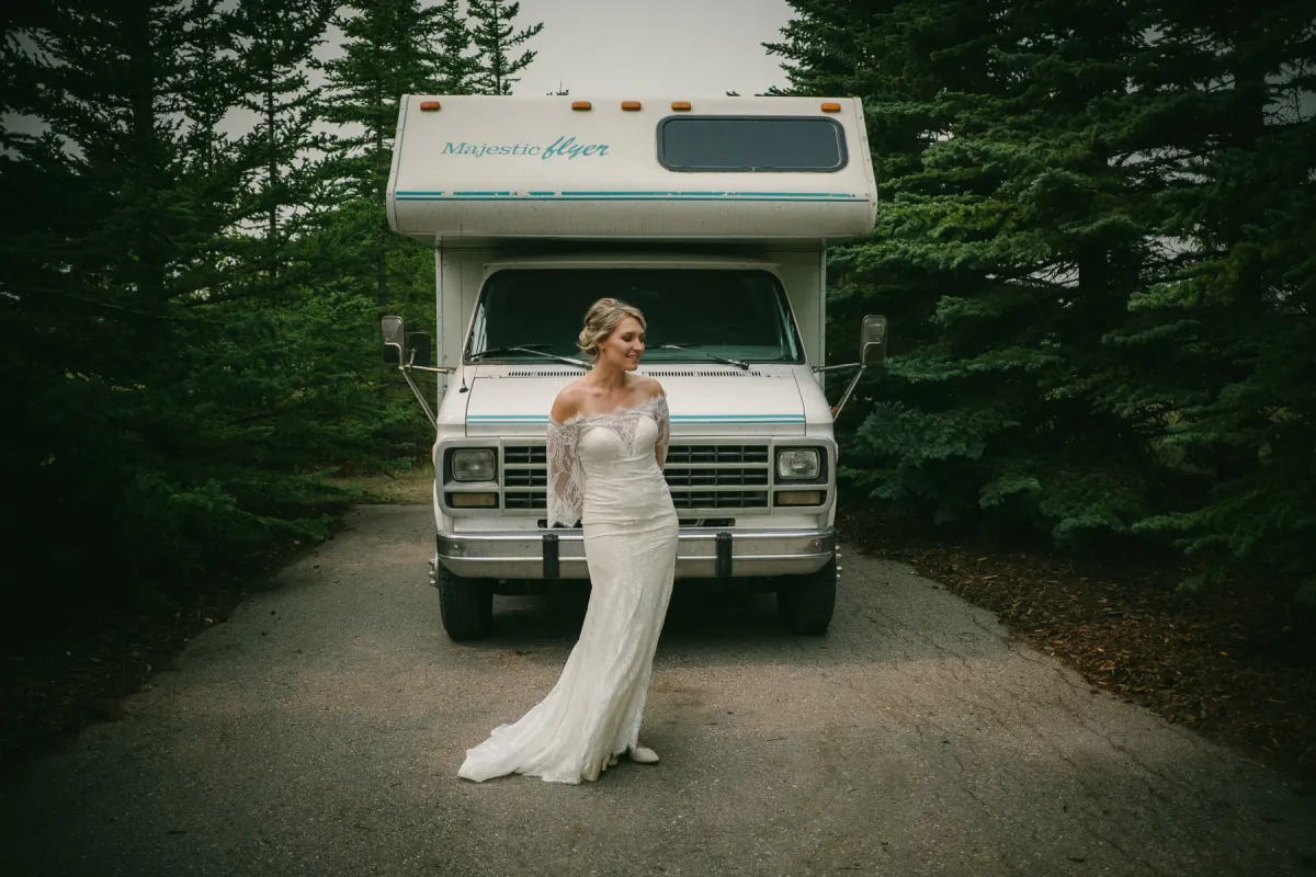 Bride in an off-shoulder lace gown standing before a white Majestic Flyer motorhome in the forest