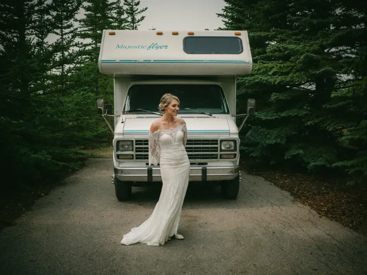 Bride in an off-shoulder lace gown standing before a white Majestic Flyer motorhome in the forest