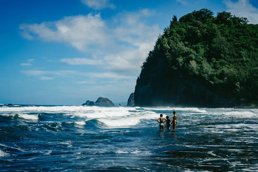 People standing in bright blue water near a black sand beach in Hawaii