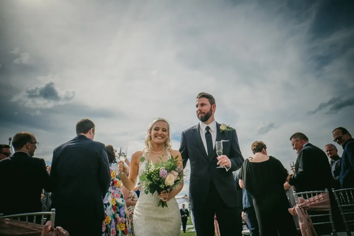 Newlyweds walking an outdoor aisle holding champagne flutes under a dramatic cloudy sky