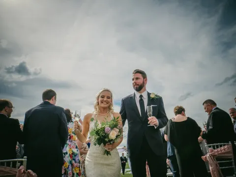Newlyweds walking an outdoor aisle holding champagne flutes under a dramatic cloudy sky
