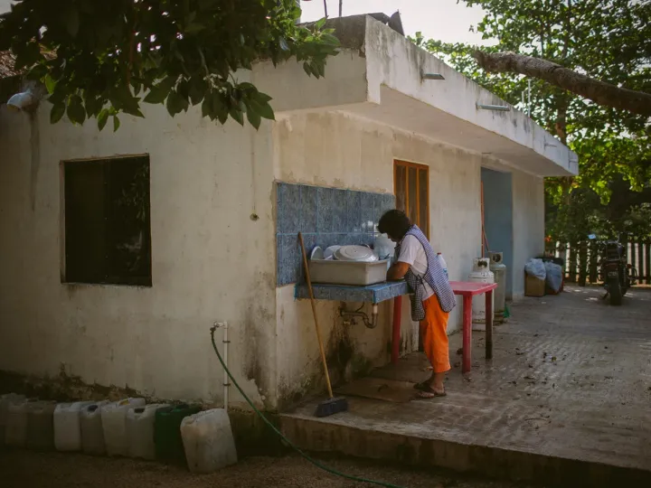 Person standing at an outdoor sink in a shaded courtyard beside a weathered building