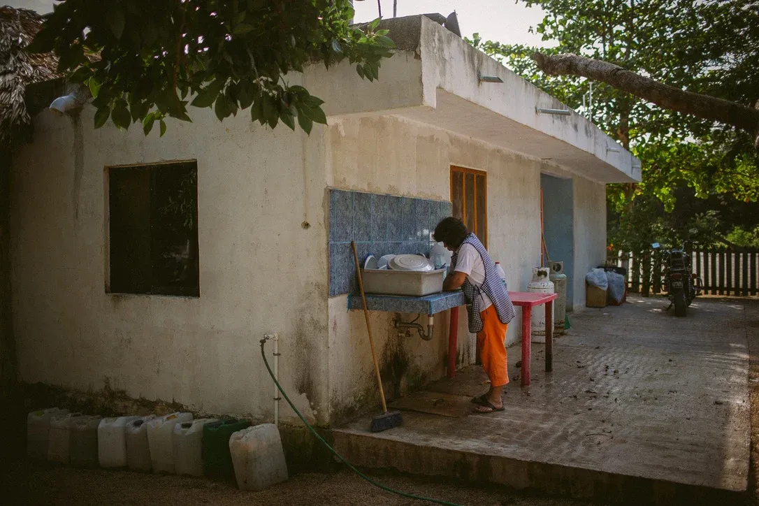 Person standing at an outdoor sink in a shaded courtyard beside a weathered building