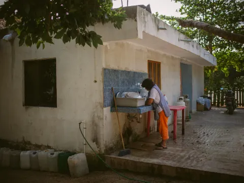 Person standing at an outdoor sink in a shaded courtyard beside a weathered building