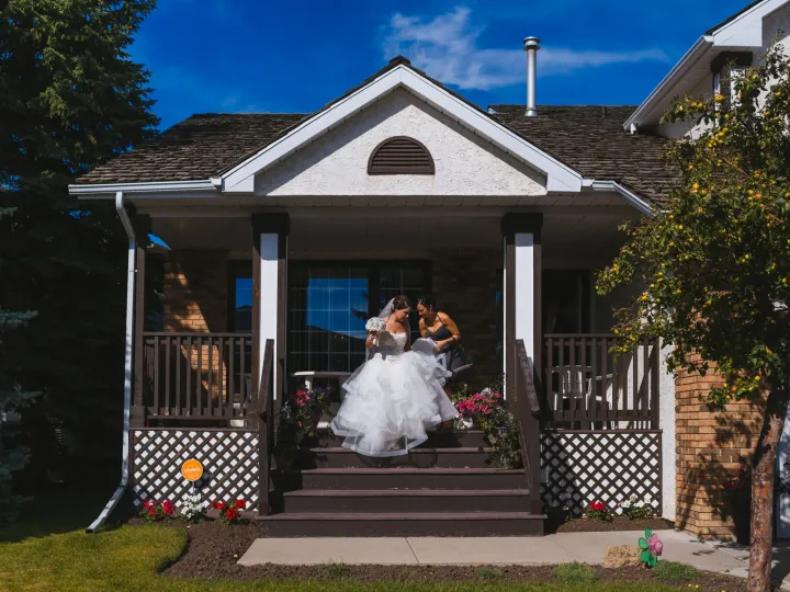 Bride in a voluminous tulle gown being helped down front porch steps of a suburban home