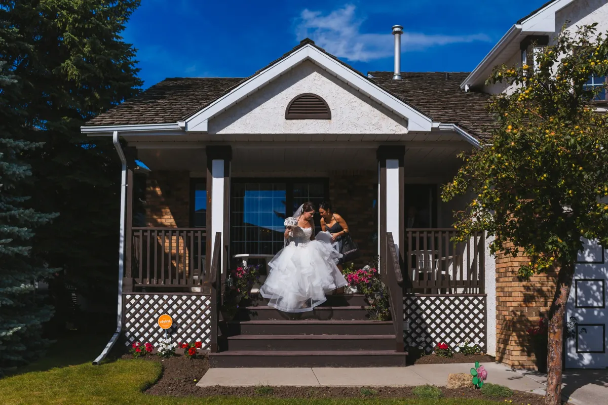 Bride in a voluminous tulle gown being helped down front porch steps of a suburban home