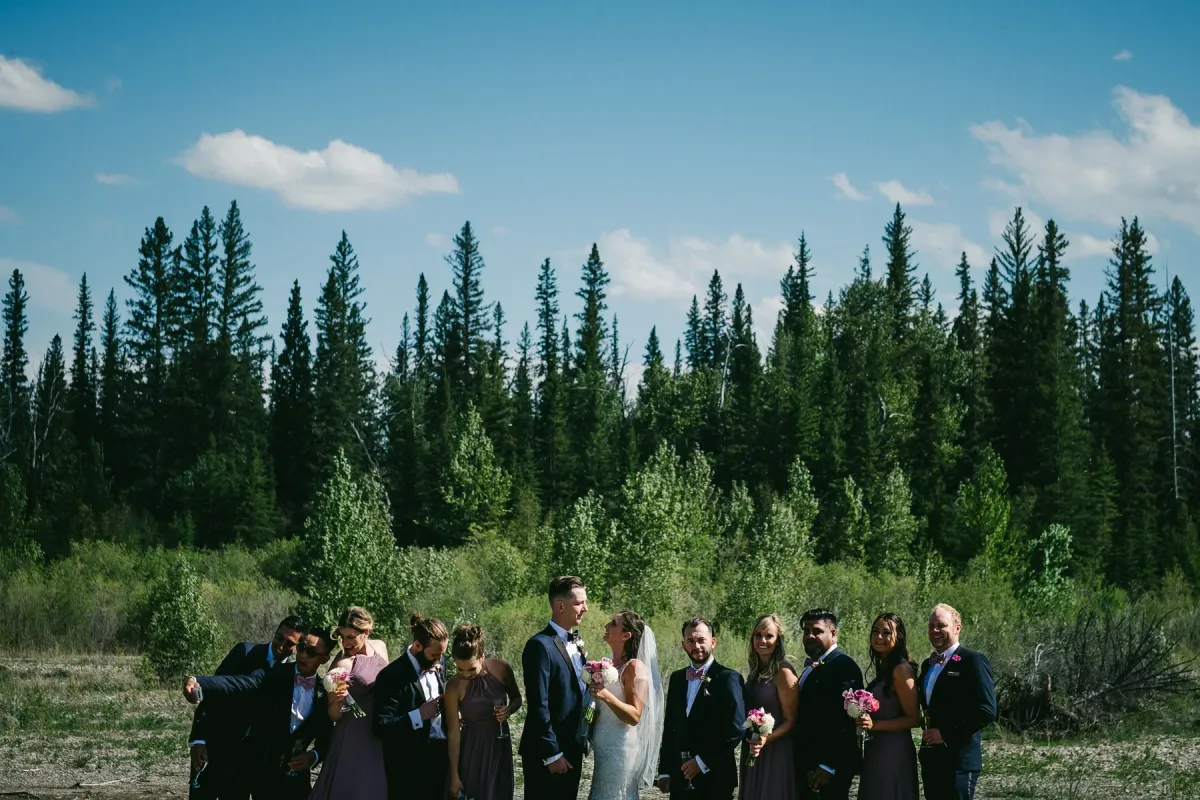 Wedding party of twelve standing in a gravel clearing before evergreen forest under a blue sky