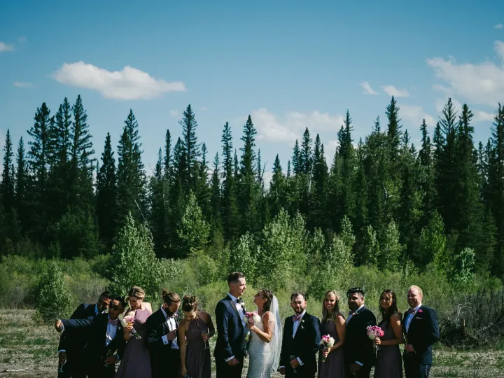 Wedding party of twelve standing in a gravel clearing before evergreen forest under a blue sky