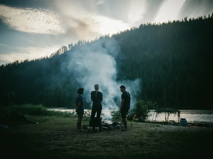 Small group gathered around a campfire beside a lake with smoke rising into evening light