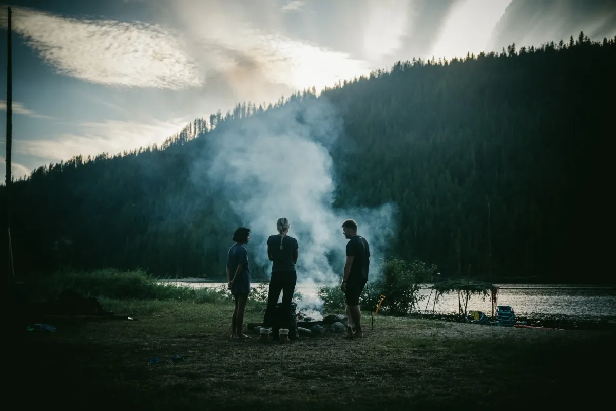 Small group gathered around a campfire beside a lake with smoke rising into evening light