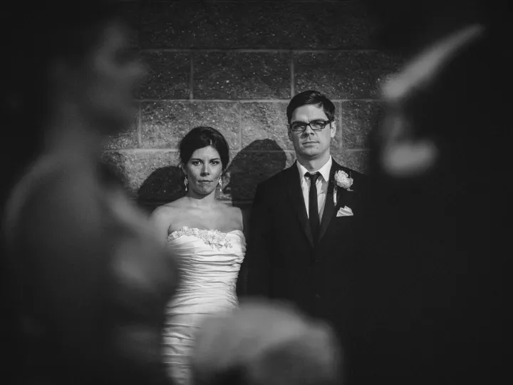 Black and white portrait of bride and groom looking at camera through blurred foreground figures