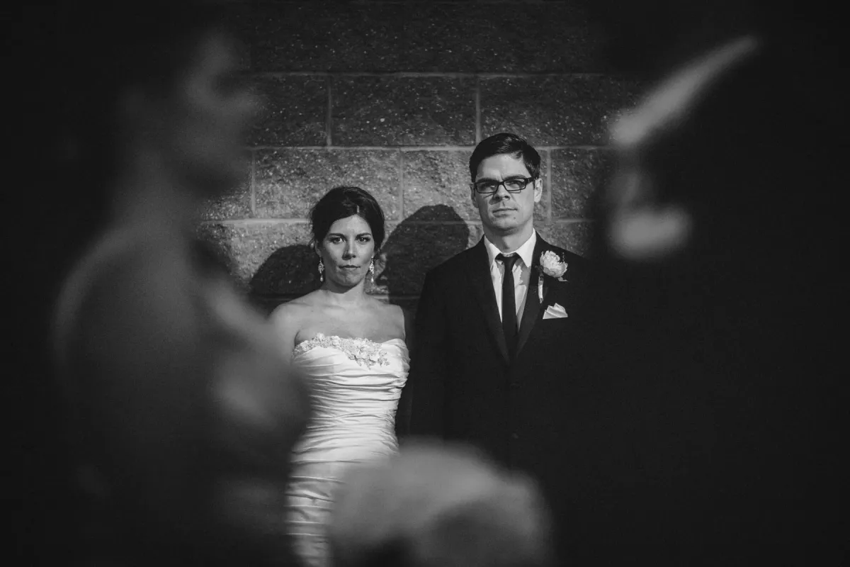 Black and white portrait of bride and groom looking at camera through blurred foreground figures