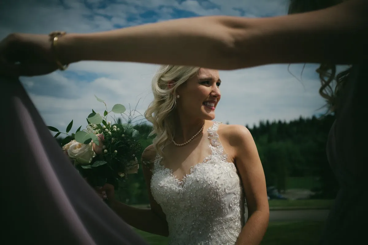 Bride in lace laughing, framed by bridesmaids' arms holding mauve fabric outdoors