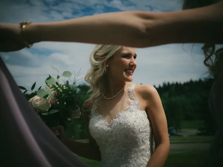 Bride in lace laughing, framed by bridesmaids' arms holding mauve fabric outdoors