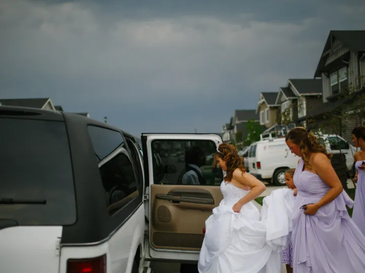 Bridesmaids in lilac dresses holding a bride's train as she enters a white SUV on a residential street
