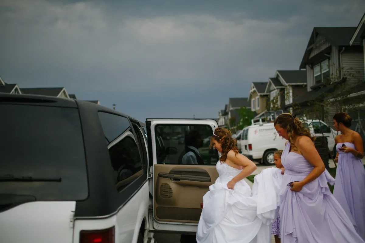 Bridesmaids in lilac dresses holding a bride's train as she enters a white SUV on a residential street