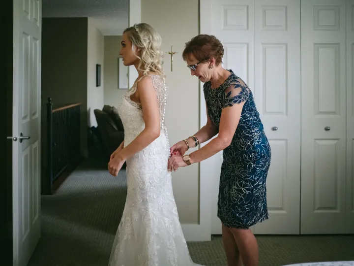 Mother in a navy dress fastening the back buttons of a bride's lace wedding gown