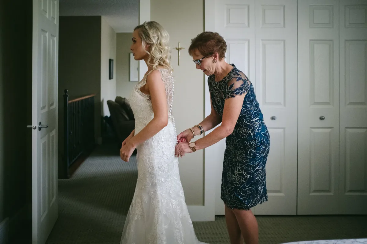 Mother in a navy dress fastening the back buttons of a bride's lace wedding gown