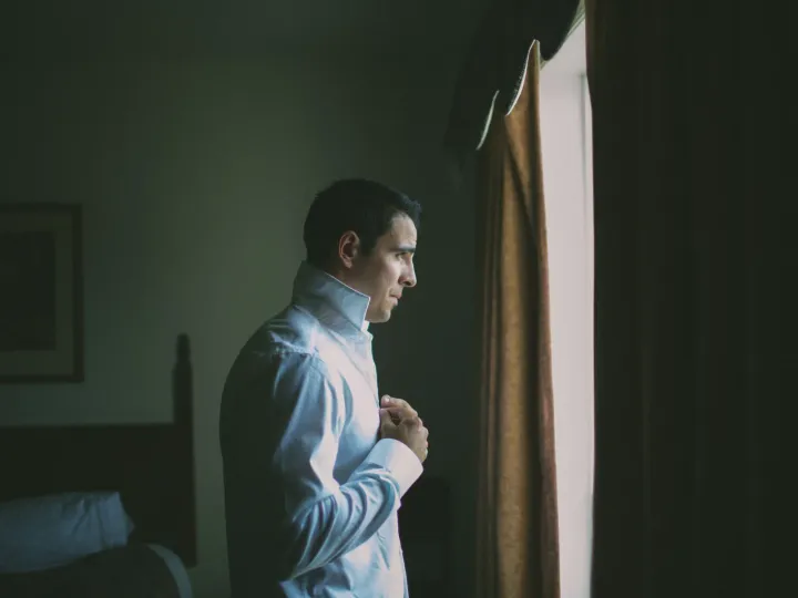 Side profile of a groom in a white dress shirt adjusting his shirt by window light