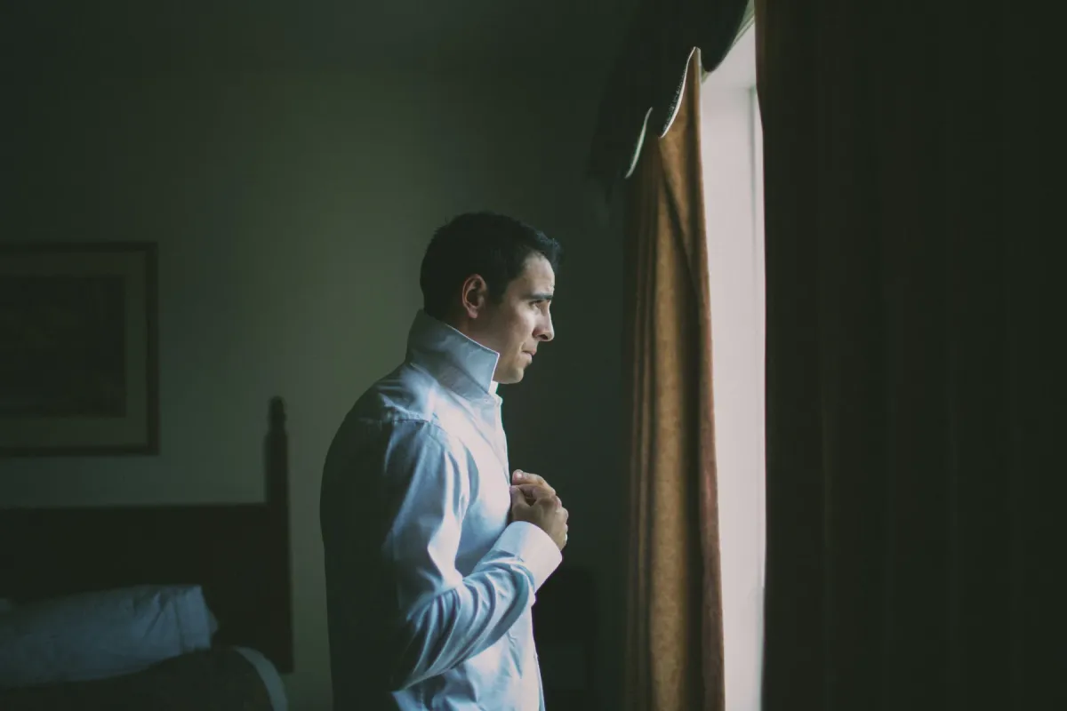 Side profile of a groom in a white dress shirt adjusting his shirt by window light
