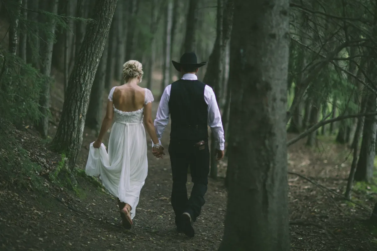 Bride and groom walking away into a dense evergreen forest, groom wearing a black cowboy hat
