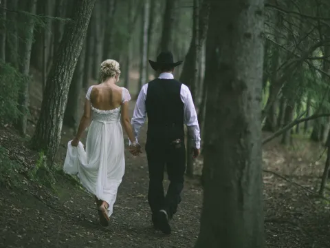 Bride and groom walking away into a dense evergreen forest, groom wearing a black cowboy hat