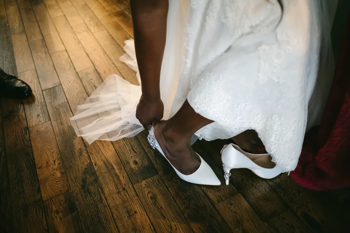 Bride leaning to fasten crystal-embellished white wedding heels on a dark wooden floor