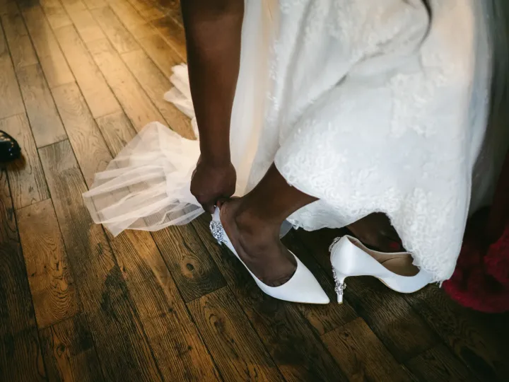 Bride leaning to fasten crystal-embellished white wedding heels on a dark wooden floor