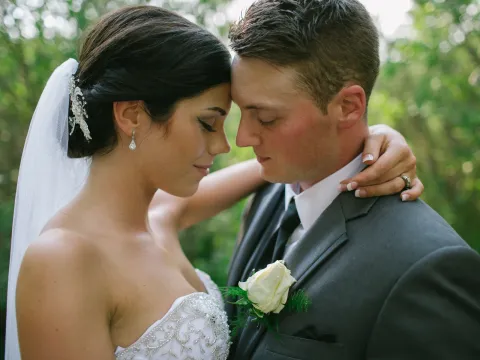 Close-up of bride and groom with foreheads touching outdoors against green foliage