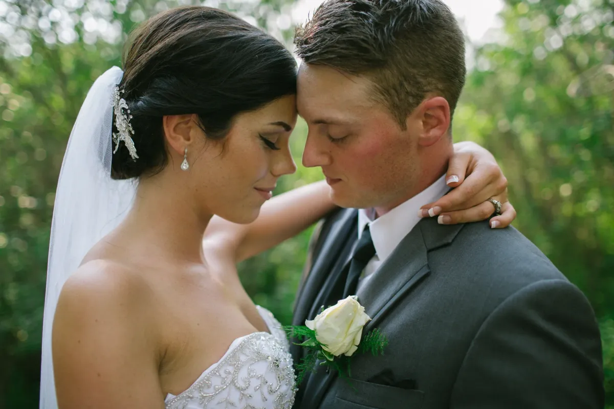 Close-up of bride and groom with foreheads touching outdoors against green foliage