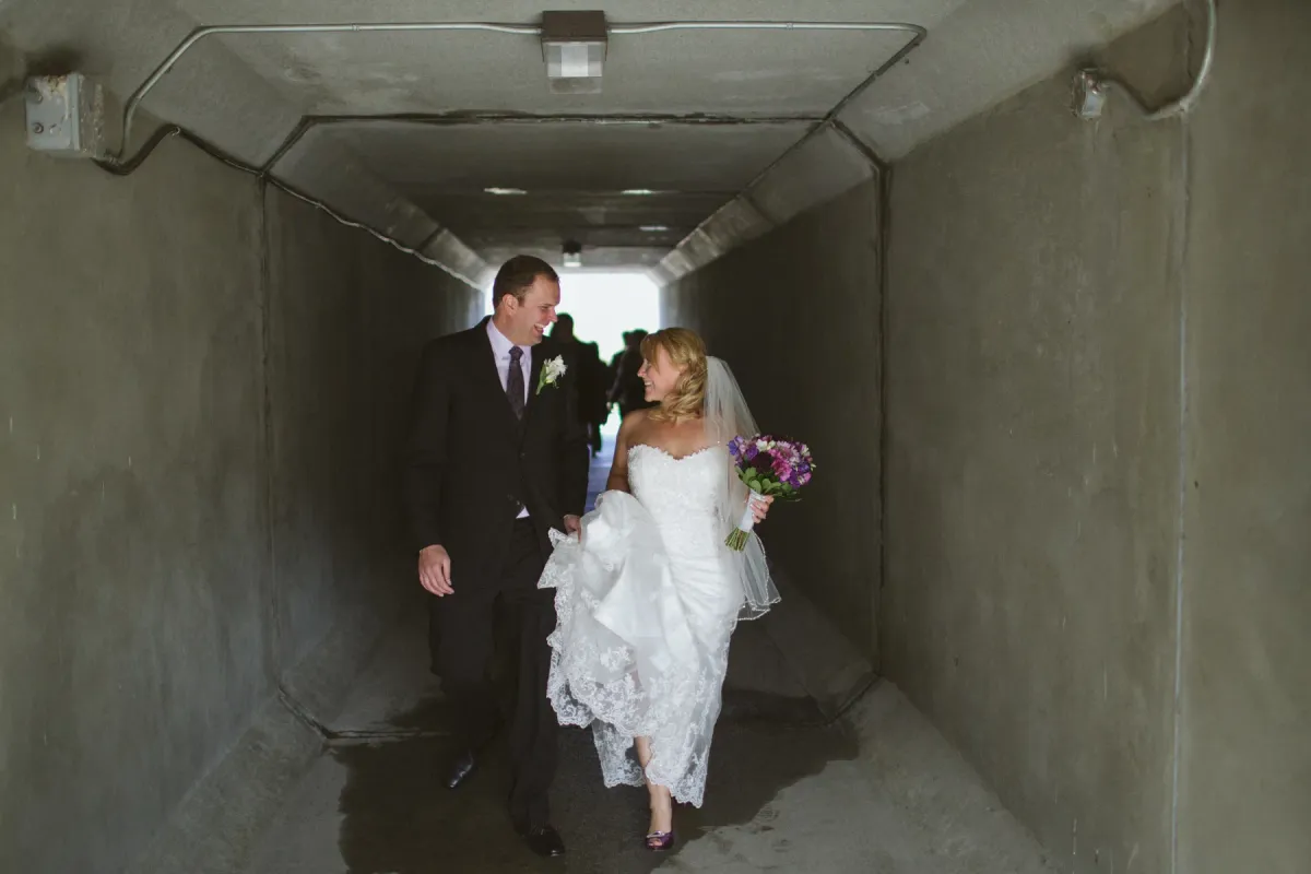 Bride and groom laughing while walking toward the camera through a concrete pedestrian tunnel
