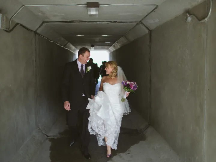 Bride and groom laughing while walking toward the camera through a concrete pedestrian tunnel