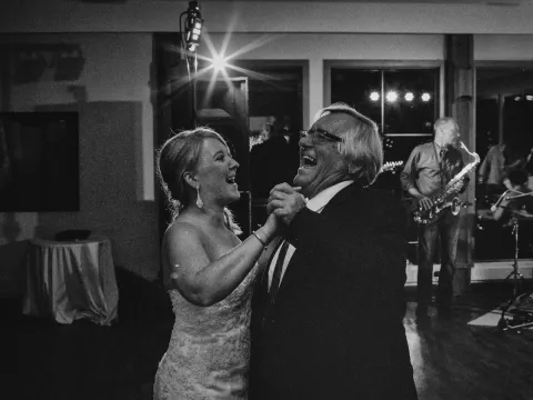 Black and white photo of a bride and older man laughing while dancing at a wedding reception