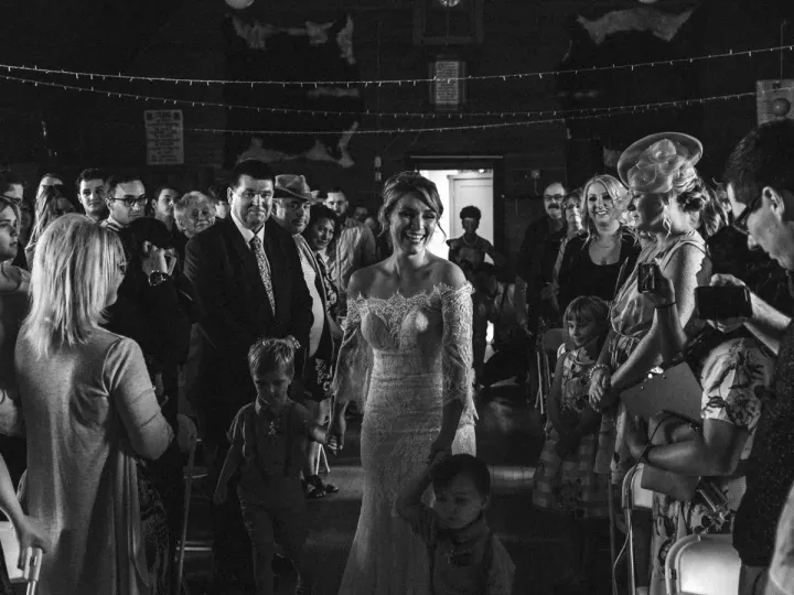 Black and white photo of a bride walking down a rustic hall aisle holding children's hands