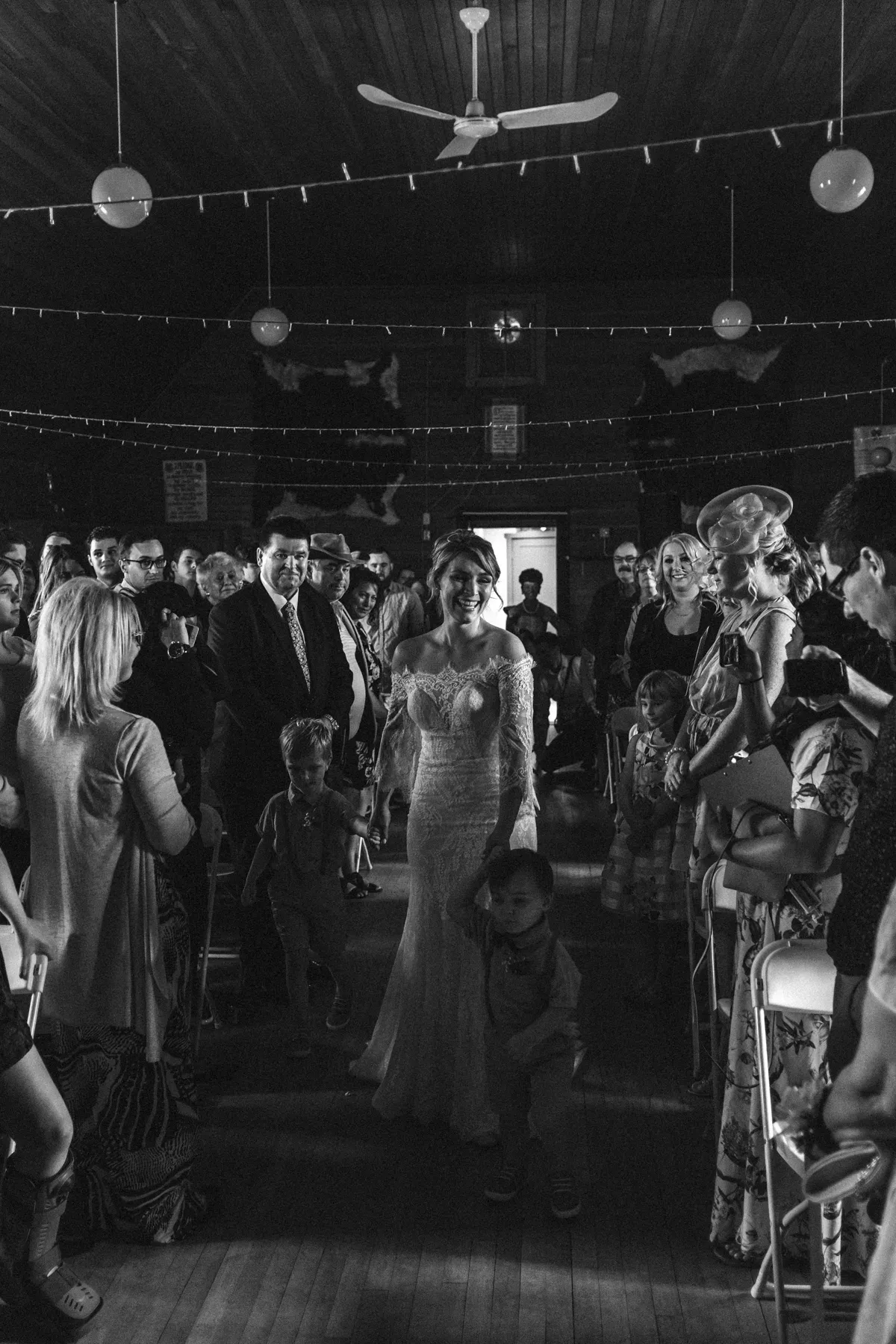 Black and white photo of a bride walking down a rustic hall aisle holding children's hands