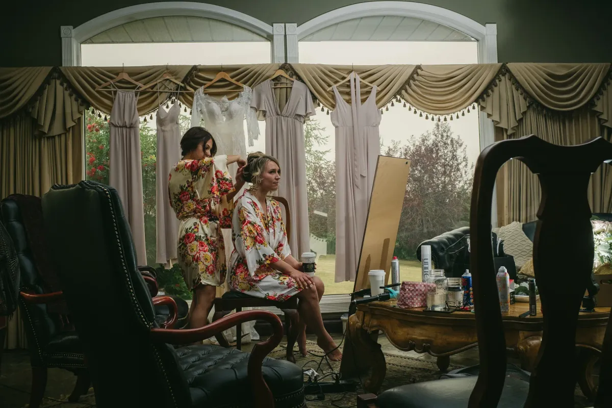 Bride in a floral robe having her hair styled while wedding dresses hang in arched windows