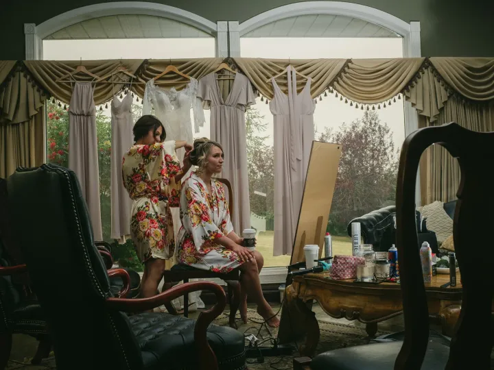 Bride in a floral robe having her hair styled while wedding dresses hang in arched windows