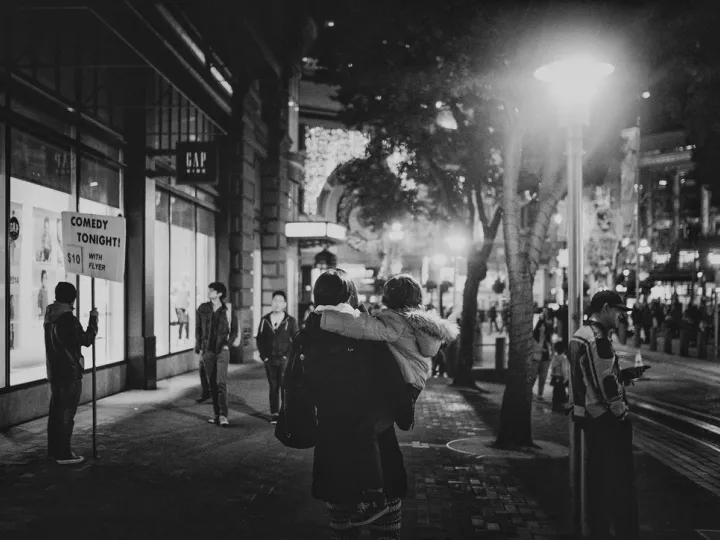 Black-and-white city street scene at night with people waiting under streetlights