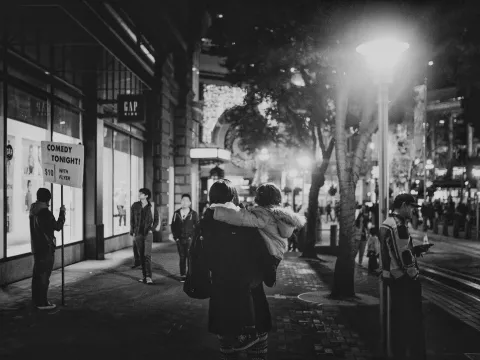Black-and-white city street scene at night with people waiting under streetlights