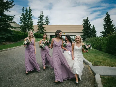 Bride and four bridesmaids in lilac dresses walking toward the camera on a paved driveway