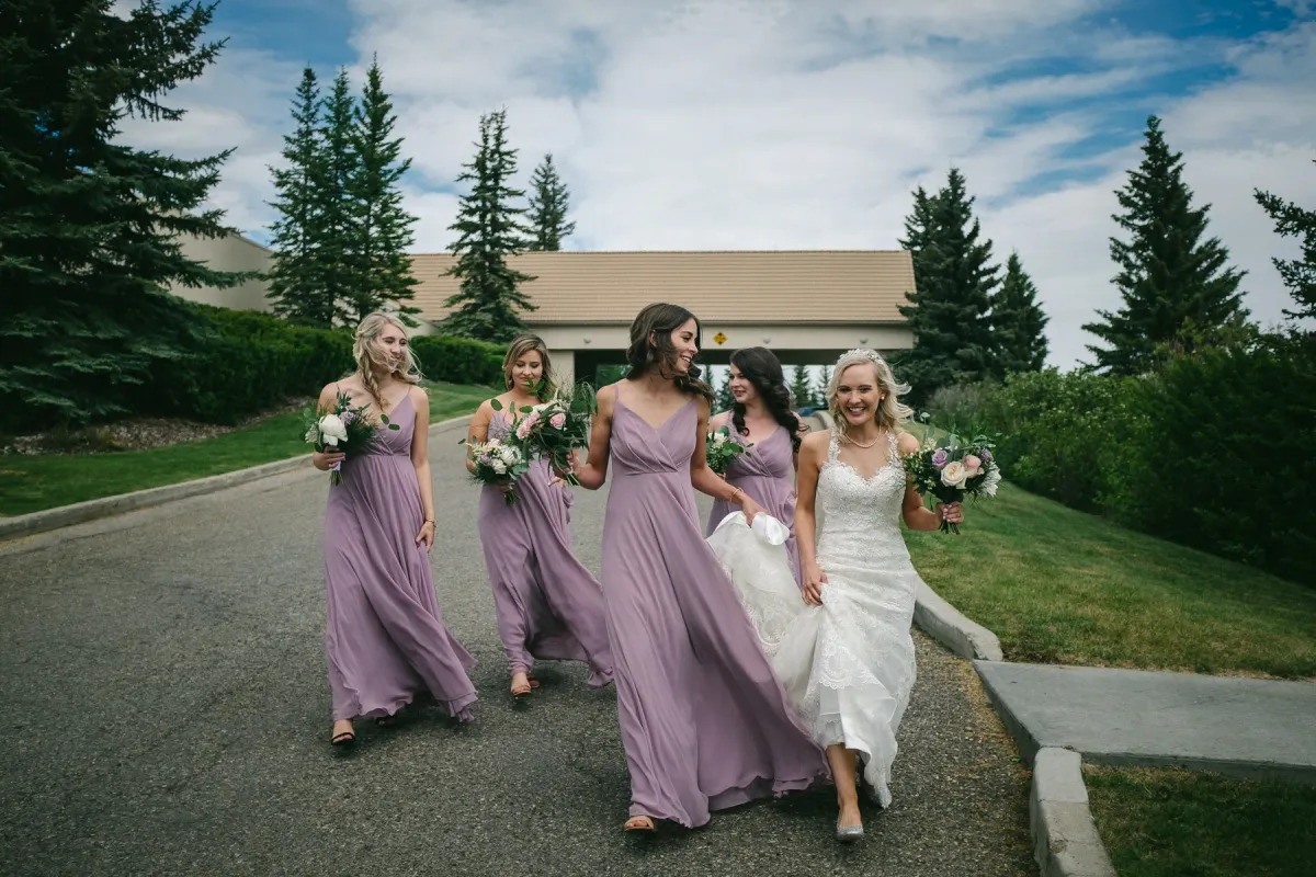 Bride and four bridesmaids in lilac dresses walking toward the camera on a paved driveway