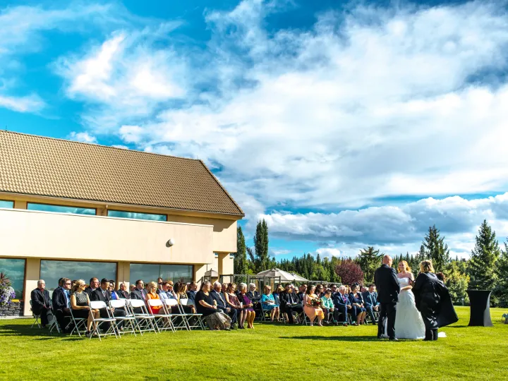 Wide view of an outdoor lawn wedding ceremony with seated guests and a large venue building