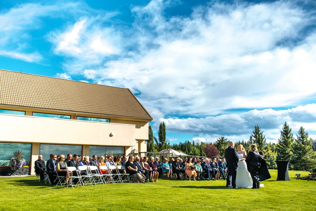 Wide view of an outdoor lawn wedding ceremony with seated guests and a large venue building