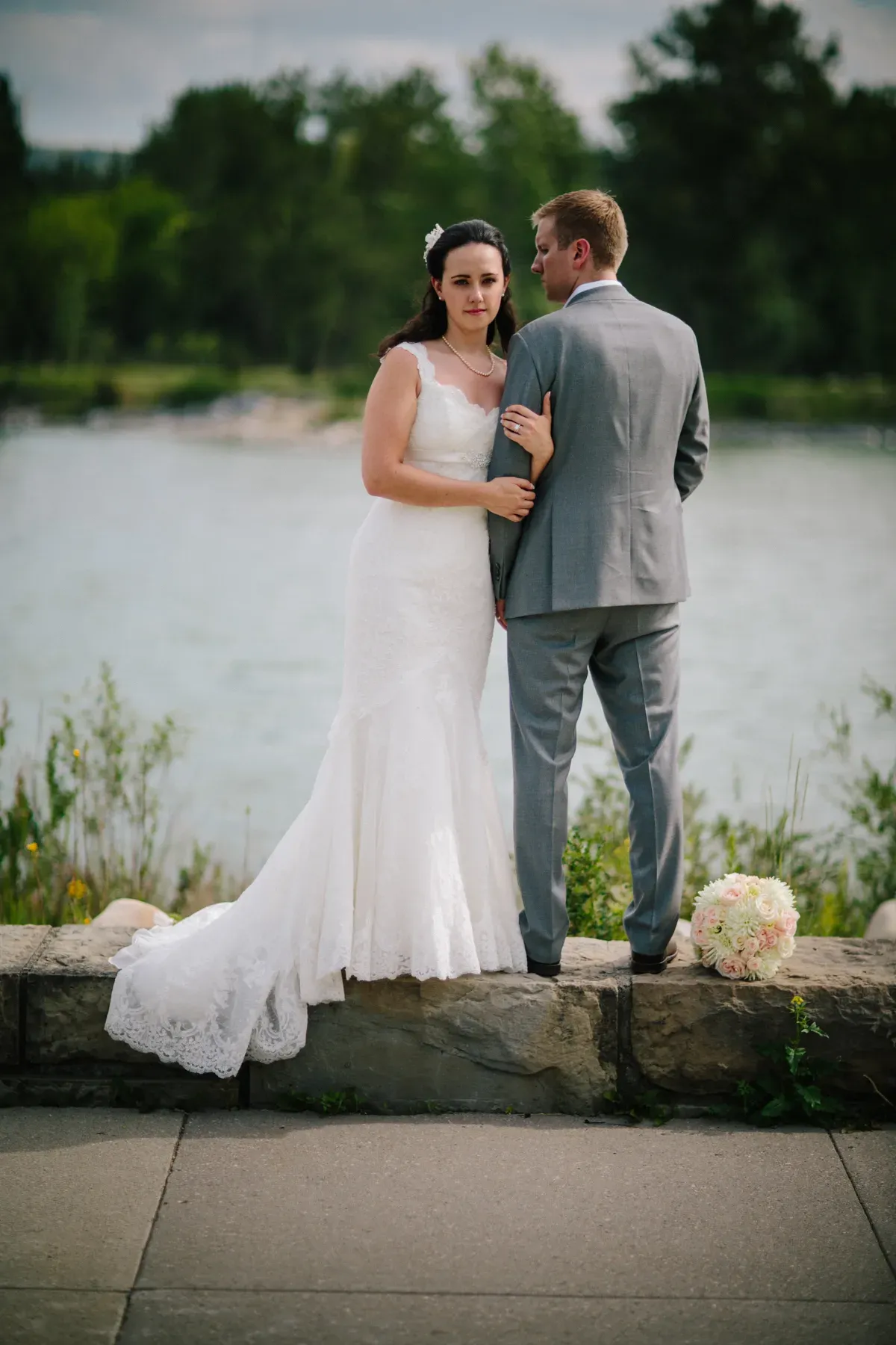 Bride leaning on a groom in a grey suit beside a river with green forest beyond