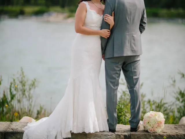 Bride leaning on a groom in a grey suit beside a river with green forest beyond